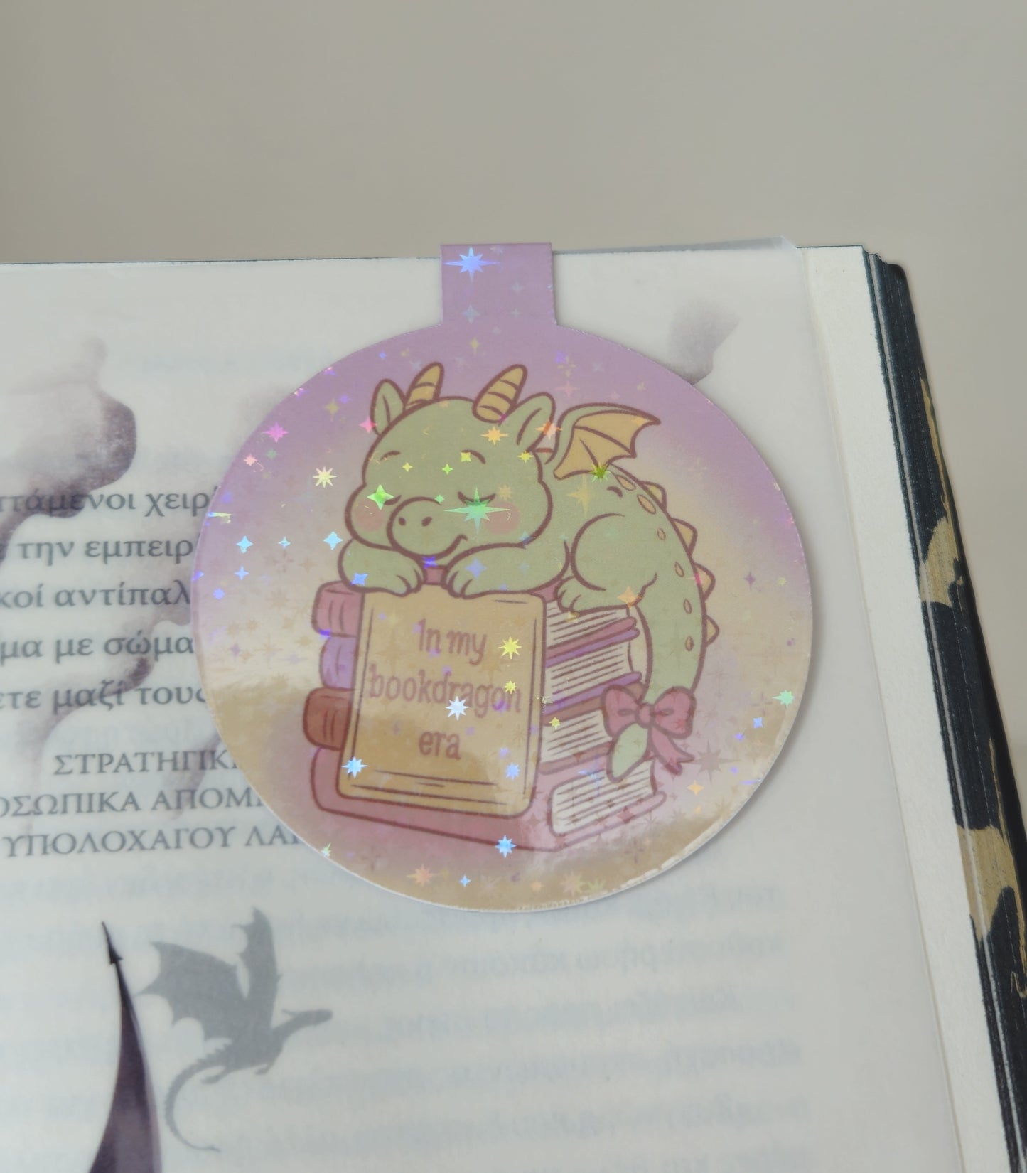 Decorative bookmark with a dragon holding a book on a white surface.